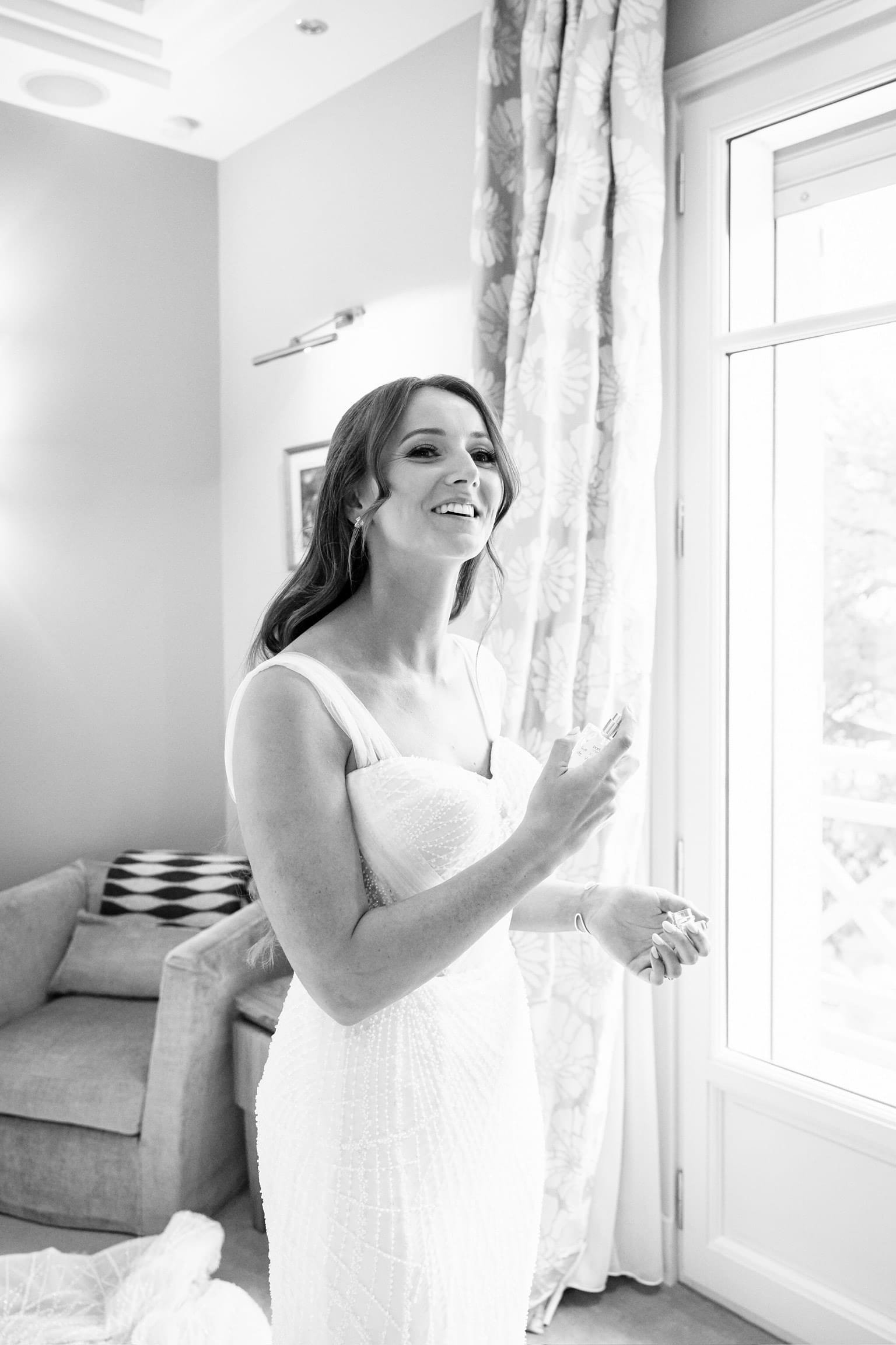 Future mariée souriante en robe blanche appliquant son parfum avant le mariage, dans une chambre lumineuse. Un moment joyeux et intime capturé dans une atmosphère élégante et sereine.