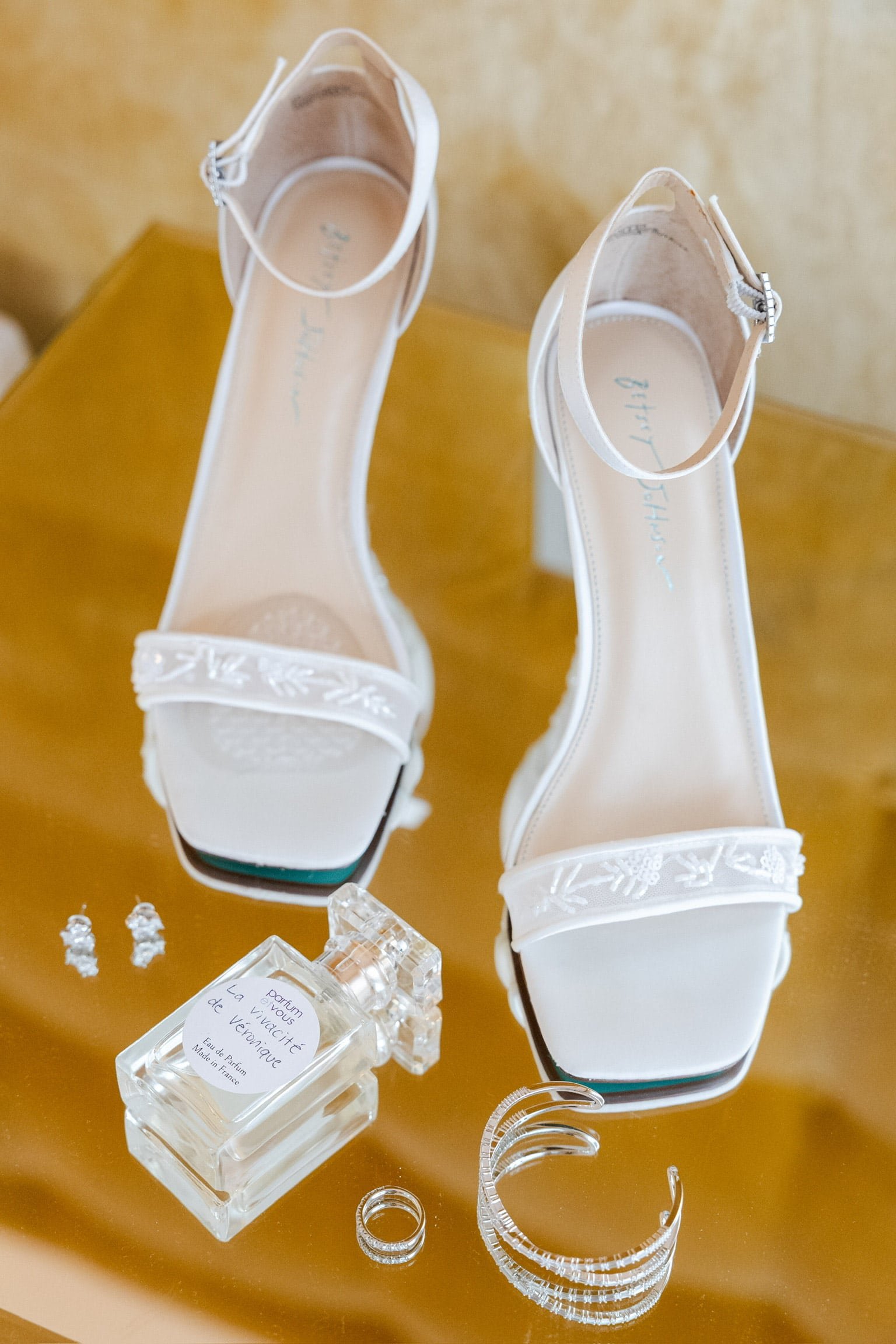 Chaussures de mariée blanches élégantes avec des détails brodés, accompagnées de parfum, boucles d'oreilles, bracelet et alliance sur une surface dorée. L'ensemble représente les accessoires raffinés de la mariée pour son grand jour